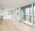 Spacious living room with large windows and sliding glass doors leading to a balcony, offering a scenic view of the city. The room has a minimalist design with light-colored hardwood floors and white walls. There is a built-in cabinet unit along one wall.