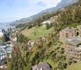 This image shows a scenic view of a lakeside town nestled in the mountains. The town features a mix of traditional and modern architecture, with a historic castle-like building and newer apartment buildings with green roofs. The town is surrounded by lush