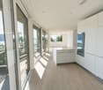 Spacious open-plan living area with large windows and sliding glass doors leading to a balcony or terrace, offering a scenic view of the lake and mountains. The room has a modern, minimalist design with white walls, hardwood floors, and built-in kitchen c