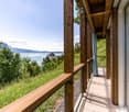 Wooden balcony with a scenic view of a lake, mountains, and a castle in the distance. The balcony has a wire mesh railing and is surrounded by lush greenery.