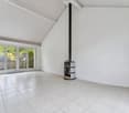 Spacious living room with large windows, white walls, and a tiled floor. The room features a wood-burning stove or fireplace in the corner.