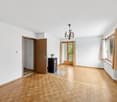 Spacious living room with hardwood parquet flooring, white walls, a wooden door, a chandelier, and a sliding glass door leading to a balcony or terrace.