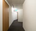 Empty hallway with white walls, wooden door, gray carpet, and an emergency exit sign