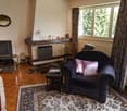 Cozy living room with a stone fireplace, hardwood floors, area rug, TV stand, and a comfortable armchair with a decorative pillow. Large windows provide natural light and a view of the surrounding trees.