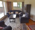 Spacious living room with large windows, balcony access, hardwood floors, patterned area rug, dark blue sofa and armchairs, wooden coffee table, and a cabinet or shelving unit