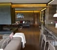 The image shows a spacious open-plan living area with a living room, dining area, and kitchen. The room features wooden beams and paneling, creating a rustic and cozy atmosphere. There is a fireplace visible, and the lighting is warm and ambient. The livi