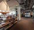 Open concept office space with high ceilings, exposed ductwork, and wooden shelving units. The space features a large sailboat model hanging from the ceiling, as well as various decorative items and books on the shelves. The office has a modern, industria