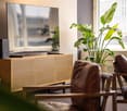 Spacious living room with large windows, a TV stand with a patterned design, a large potted plant, and a leather sofa. The room has a cozy and modern feel.