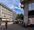 urban setting with modern buildings, people walking, cafes with outdoor seating, clock on building, greenery