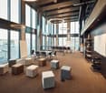 A large open room with carpeted floor, several tables, chairs, and circular ottomans, large windows with city view