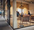 Modern office space with wooden walls, glass partitions, open floor plan, chairs and tables, glass doors, whiteboards, and monitors.