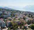Aerial view of a city nestled in a mountainous region, with a large body of water in the background. The city consists of a mix of high-rise apartment buildings, smaller residential structures, and some commercial buildings. The landscape is dotted with t