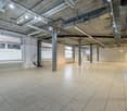 Large open commercial space with concrete ceiling, exposed pipes and ductwork, tile flooring, and windows along one wall. The space appears to be a warehouse or office area with a projector screen visible.
