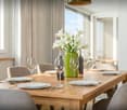 Dining room with wooden table, chairs, white plates, wine glasses, green vase with flowers, wine bottles, cutlery, and windows