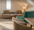living room with beige sofa, green armchair, wooden table, round rug, wooden floor, beige walls, large window