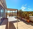 A balcony with glass walls, a bench, a table, potted plants, and a beautiful view of the mountains.