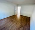 Empty room with wooden floor, white walls, open door leading to another room