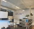 Empty room with white walls, grey ceiling, a tripod with camera, several electronic equipment on a table including a mixing console, and text that reads 'HiFLOW TV' on the wall.