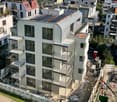 Aerial view of a modern multi-story apartment building with balconies, solar panels on the roof, and construction site nearby.