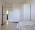 Empty bedroom with white walls and white tiled floor, bed with white bedsheet and white pillow, white wardrobe, and a chest of drawers.