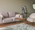 The image shows a cozy living room with a gray sofa, a round coffee table with a chess set, and a comfortable armchair. The room has a neutral color scheme with beige walls and hardwood floors. A round mirror and a framed artwork add visual interest to th