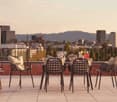 Outdoor seating area with wicker chairs and table, overlooking a city skyline with high-rise buildings and mountains in the background
