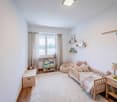 Child's bedroom with white walls, wooden floor, white rug, bed, shelves, lamp, storage box, bean bag, toys, books, picture frames, window with curtains