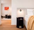 white walls, wooden floor, wall lamp, double bed with white pillows, dark nightstand, artwork, curtain