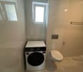 Small bathroom with white tiles, washing machine, toilet, and a window