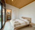 A modern bedroom featuring a double bed with clean, crisp white bedding, wooden side table, wooden ceiling, glass sliding door, adjacent bathroom with sink, small wooden cabinet, and a mirror.
