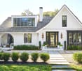 Two-story white house with a gable roof, black windows, and a covered front porch. The house has a detached garage and a chimney, indicating a fireplace. The landscaping includes shrubs, potted plants, and a stone pathway leading to the front entrance.