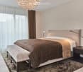 Spacious bedroom with large bed, bedside tables, lamps, chandelier, carpet, white and brown drapes, bed bench, clean white walls.