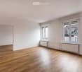 Empty room with white walls, wooden floor, two windows with built-in white radiators, door leading to another room