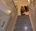 Stairs with wooden and gray steps, white walls, gray handrail, white door at top, ceiling light, clothes hanging on wall.