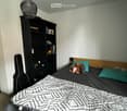 Queen size bed with dark gray bedding, dark book shelves with books and decorative items, black guitar case, light gray walls