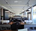 Open floor plan office, large desks, computer monitors, shelves, black hanging lamps, plants, open windows, exterior glass wall