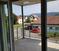 An exterior view through a sliding glass door onto a balcony with a metal railing and a red car parked in front of another house.