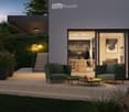Modern living room with large windows, wooden floor, sofa, outdoor lounge chairs, coffee tables, decorative lights, kitchen area visible through the windows.
