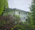House. A modern, single-story house with a flat roof and light green exterior, surrounded by lush greenery and trees.