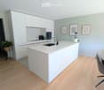 modern kitchen with white cabinets, island countertop, wooden floor, framed pictures on wall, potted plant, modern chair