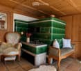 Cozy room with wooden walls and ceiling, green tiled stove, floral-patterned chair, small table with lamp, and decorative items.