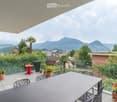 Balcony / Terrace. A modern balcony with a glass railing, offering a scenic mountain view. It features a large dining table with chairs, several potted plants, and a small red side table.