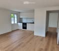 Unfurnished Room. Empty room with light wooden flooring, white walls, and a small kitchen area with white cabinets, a sink, and an oven. A doorway leads to another room.