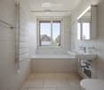 Modern bathroom with a bathtub, sink, toilet, mirror, and showerhead. The room features large windows with a view of the outdoors.