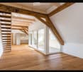 Bright, open loft space with wooden floors, exposed wooden beams, and a staircase leading to an upper level. Features large glass doors and white walls.