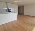 Empty room with light wooden flooring, white walls, and a modern kitchen area with white cabinets and a marble countertop.
