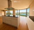 Modern kitchen with light-colored cabinets, a central island, and expansive windows offering a scenic view of a lake and landscape.