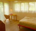 Bright bedroom with wooden flooring, large windows with sheer curtains, a bed with a green mattress and floral duvet, a wicker chair, a small wooden table with a plant, and a wall-mounted lamp.