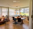 Bright, open living and dining area with wooden flooring, large windows, and modern furniture including a sofa, dining table, and pendant lights.