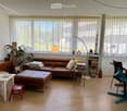 Bright living room with large windows, leather sofa, ottoman, coffee table, floor lamp, guitar, and various decorative items.